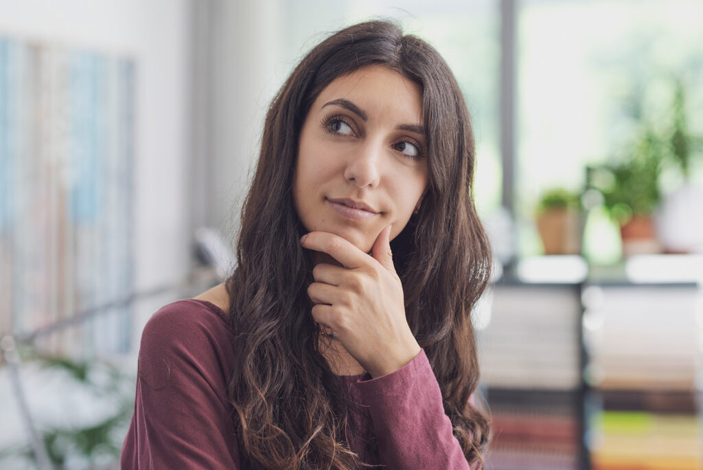 A young woman with long, wavy brown hair rests her chin thoughtfully on her fingers, gazing off to the side in an indoor setting