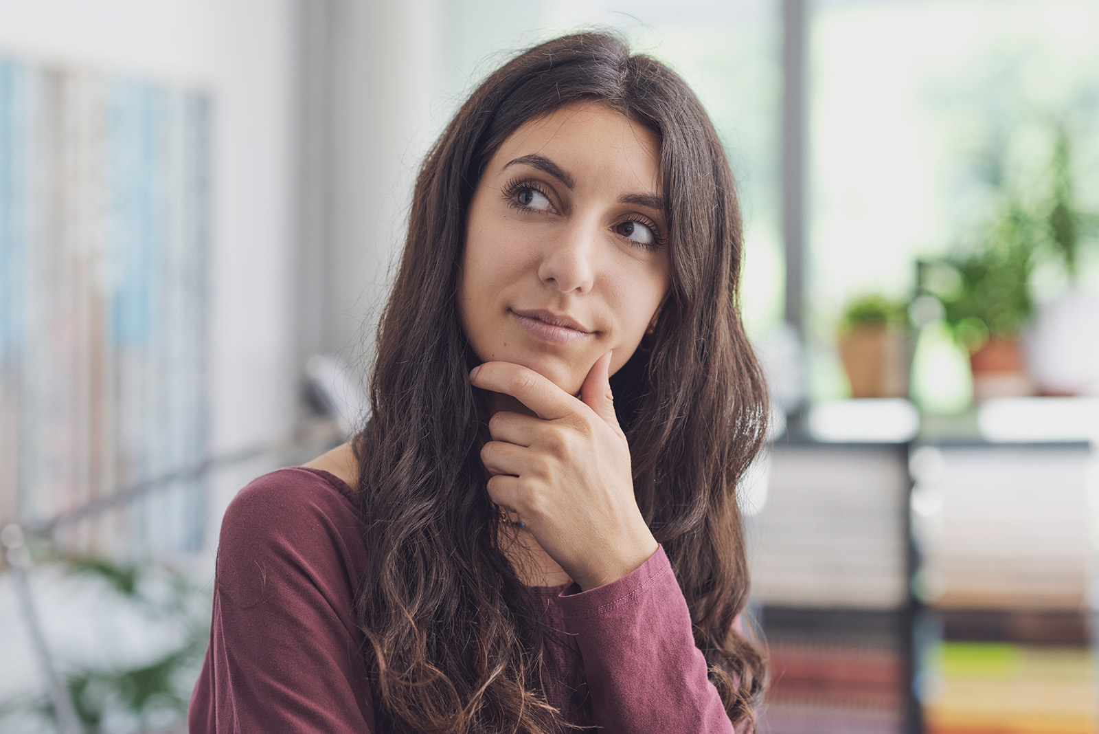 A young woman with long, wavy brown hair rests her chin thoughtfully on her fingers, gazing off to the side in an indoor setting
